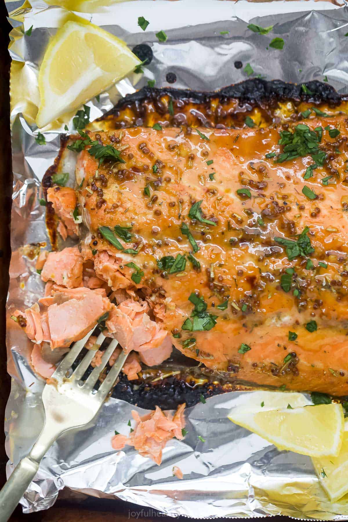 Dijon maple glazed salmon is a low-calorie, high-protein dinner that's done in just 15 minutes! The tangy-sweet glaze caramelizes the skin while the inside bakes to tender, flaky perfection. A baked salmon fillet on top of a foil-lined baking sheet with a fork flaking some meat off of the fillet.