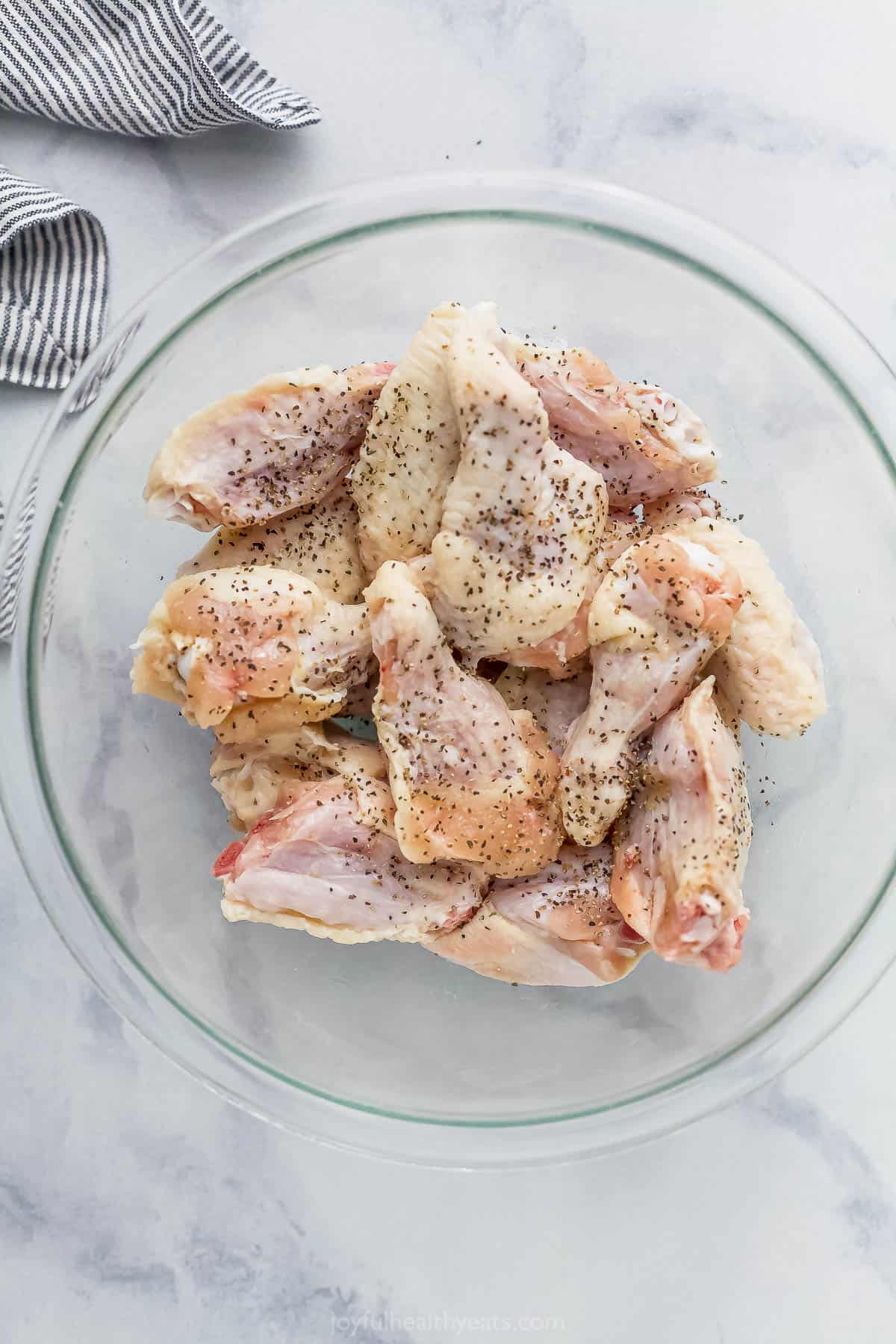 A large glass bowl full of raw chicken wings seasoned with salt and pepper