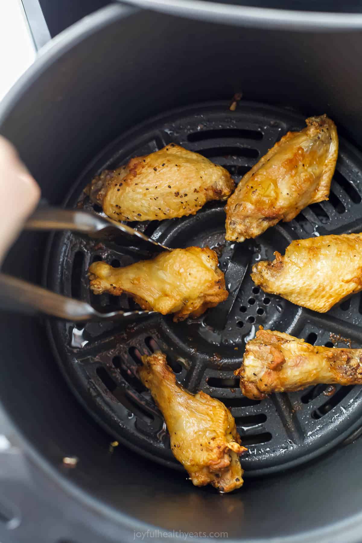 Partially cooked wings in an Air Fryer basket being flipped with a pair of metal tongs