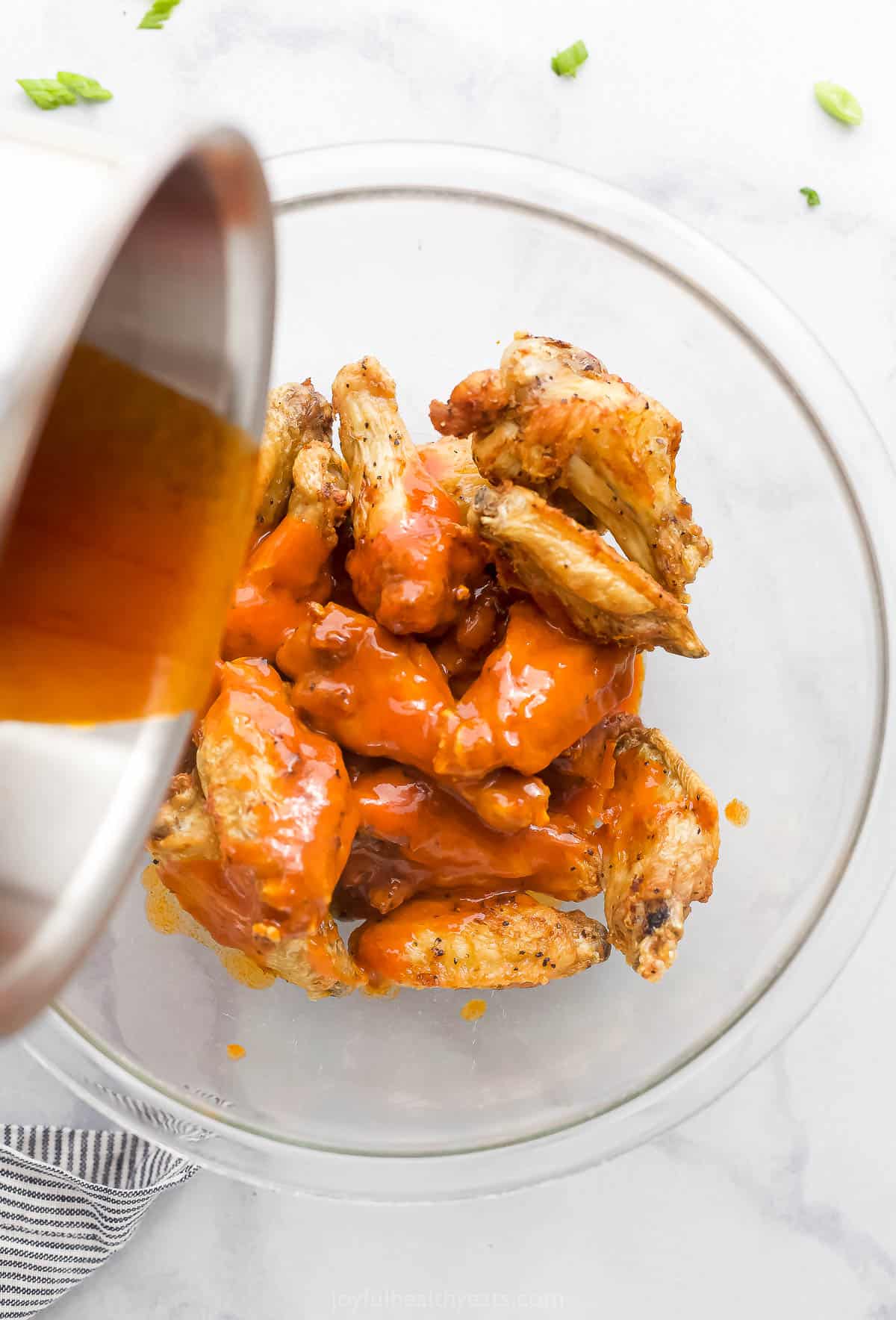 A bowl of buffalo sauce being poured into a bowl filled with air-fried chicken wings