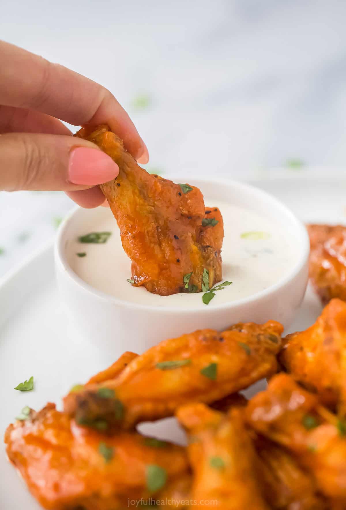 A hand dipping a chicken wing into a small bowl of ranch dressing