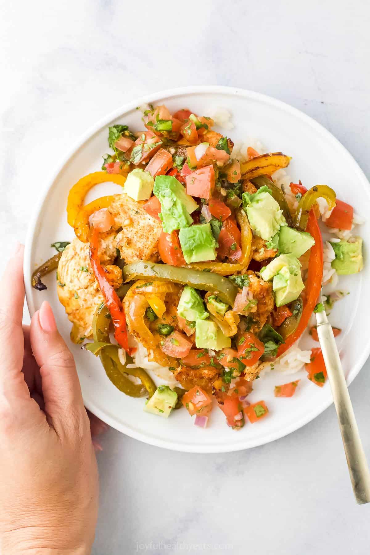 A hand holding a white plate with sheet pan chicken fajitas