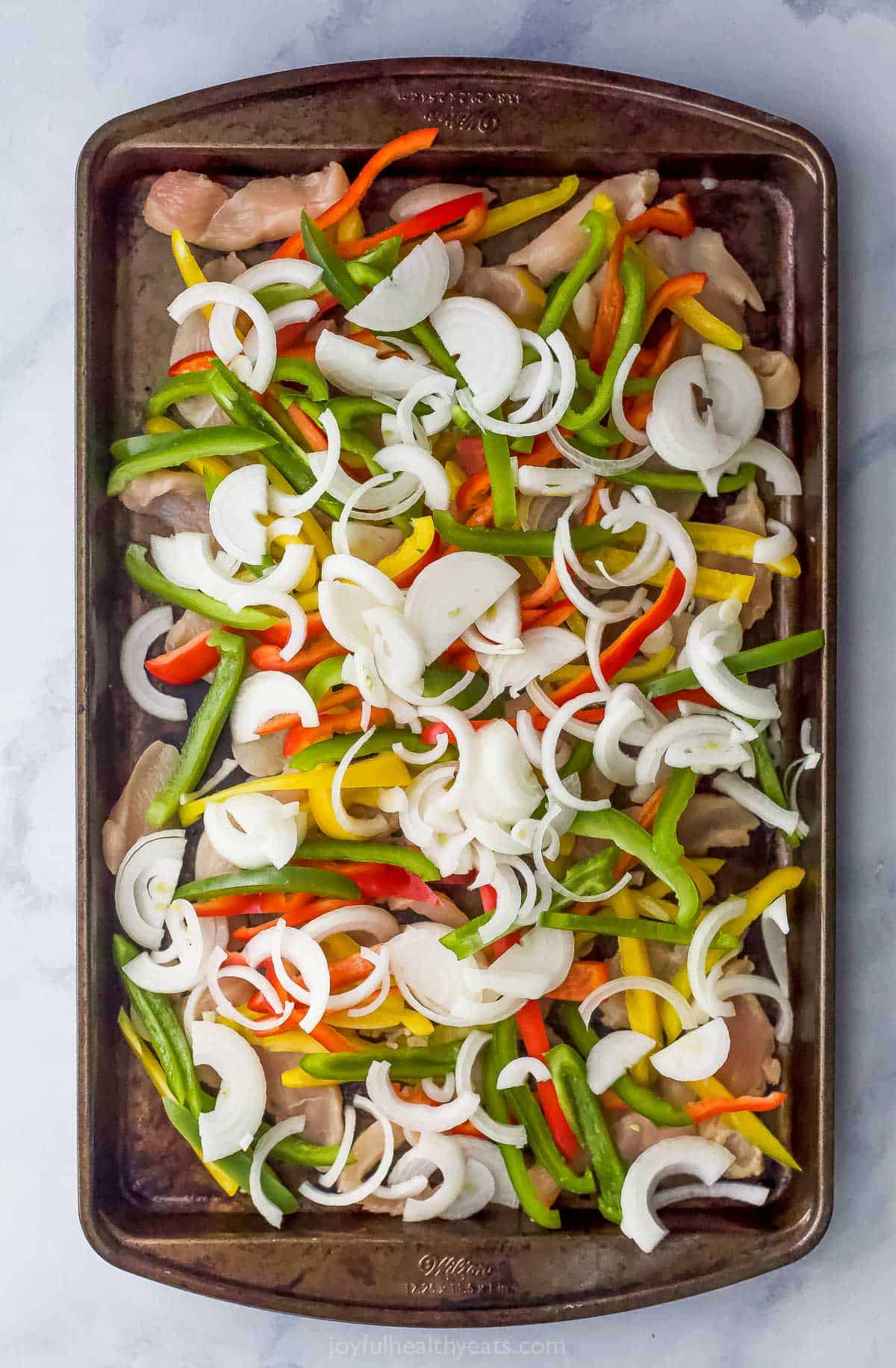 Onions on top of peppers on topped of chicken breast slices on a baking sheet