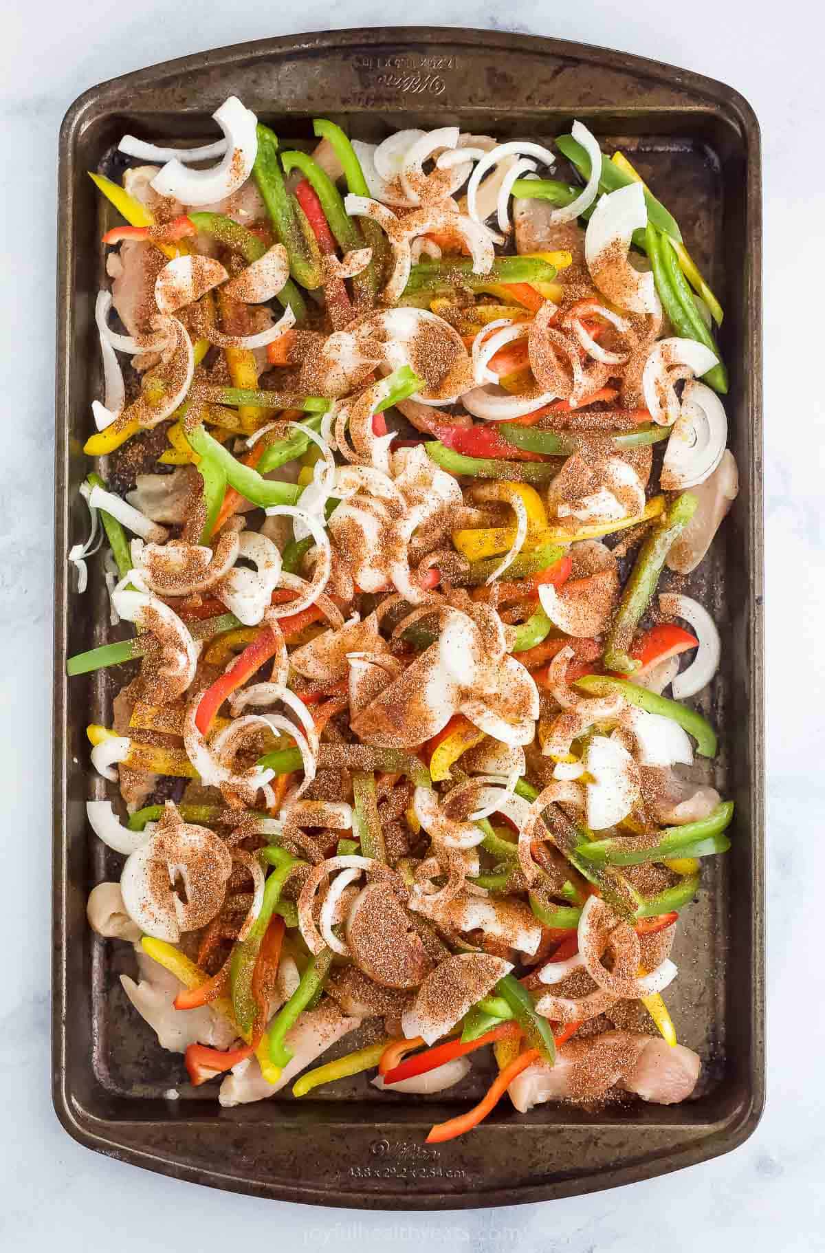Peppers, onions, and chicken breast slices mixed with fajita seasoning on a baking sheet