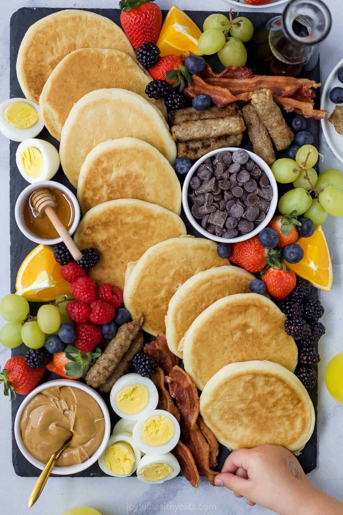 A charcuterie board full of pancakes, fruits, meats and condiments with a hand reaching for a piece of bacon
