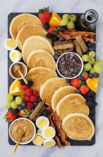 A charcuterie board containing a bunch of breakfast food including pancakes and maple syrup