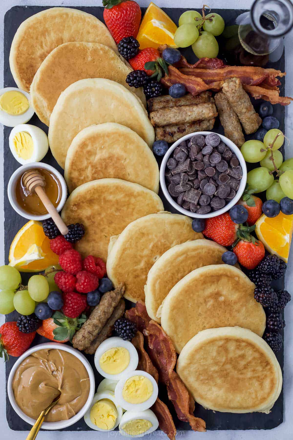 A black serving platter with pancakes, hard-boiled eggs, berries, sausage and more breakfast food on top of it