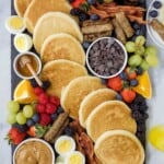 A brunch buffet board on top of a marble surface with a hand reaching for a strip of bacon