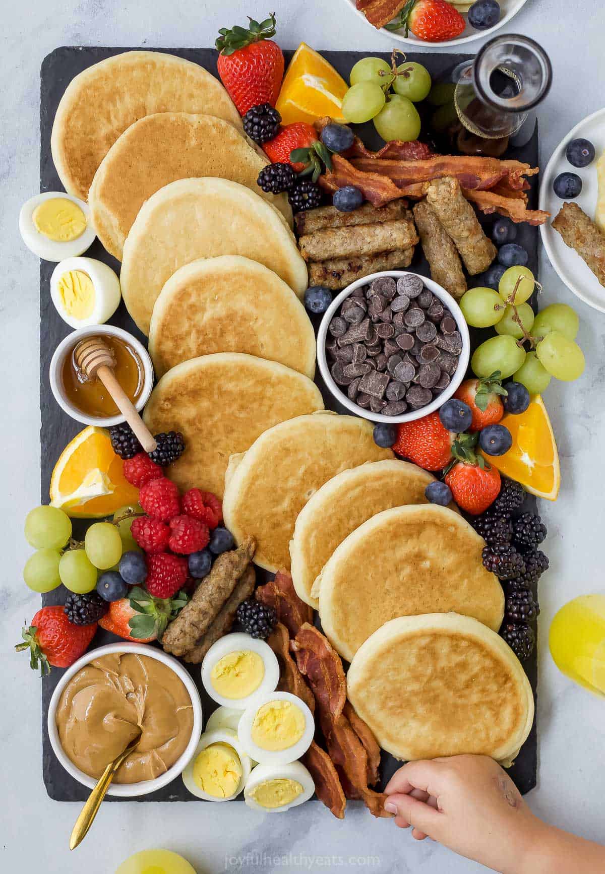 A brunch buffet board on top of a marble surface with a hand reaching for a strip of bacon