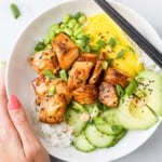 A hand holding a salmon poke bowl with a pair of chopsticks resting on the rim