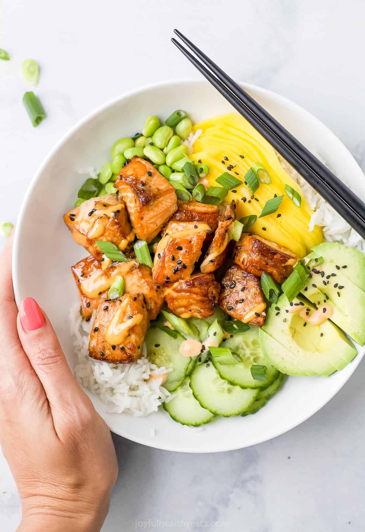 A hand holding a salmon poke bowl with a pair of chopsticks resting on the rim