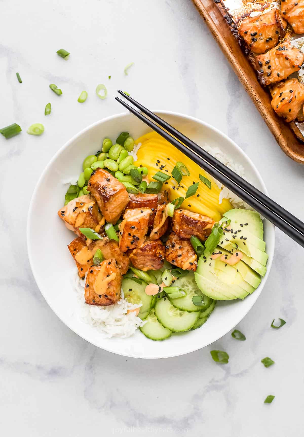 A salmon poke bowl on a marble countertop beside a pan full of roasted salmon