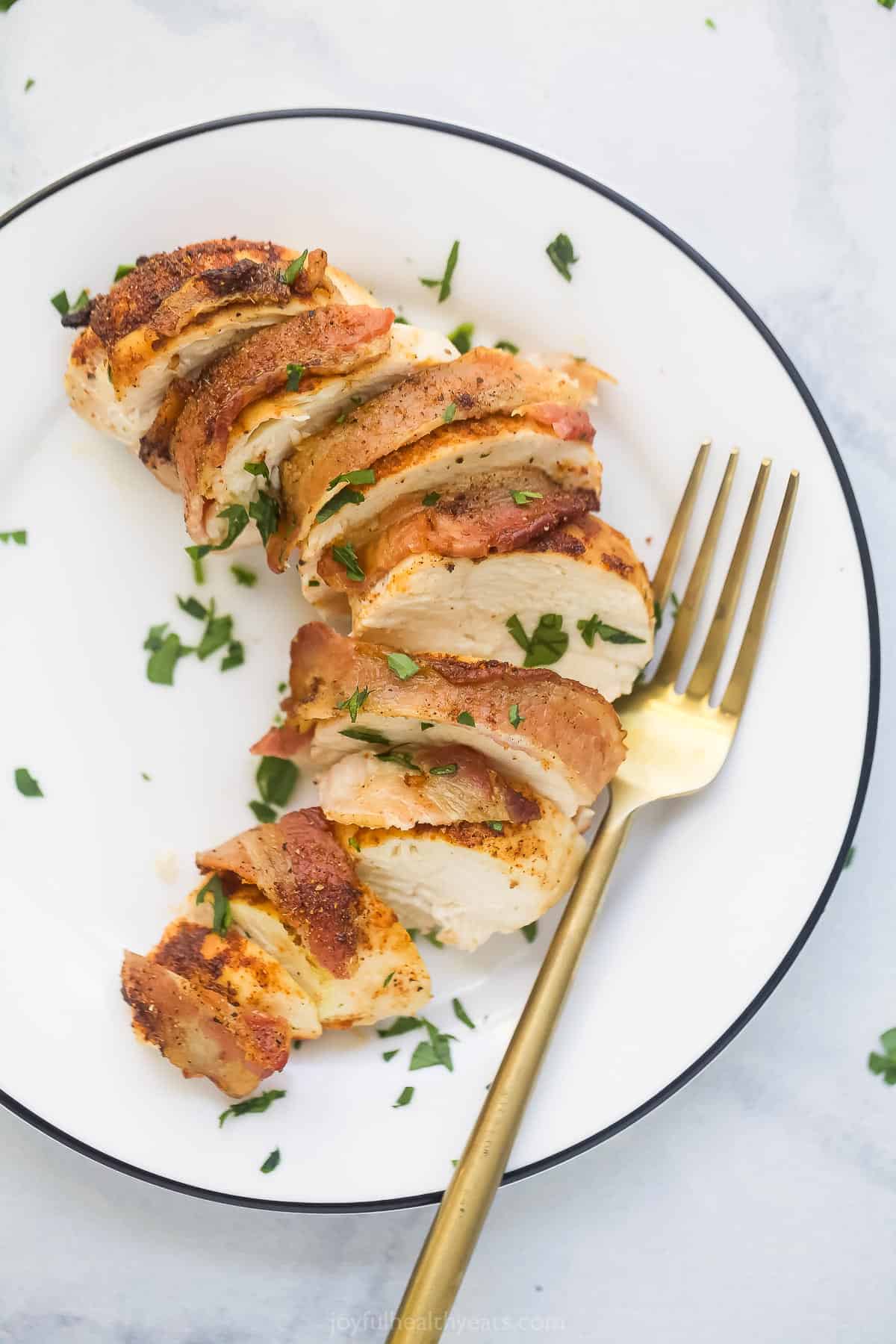 Sliced bacon wrapped chicken breast on a white plate with a gold fork