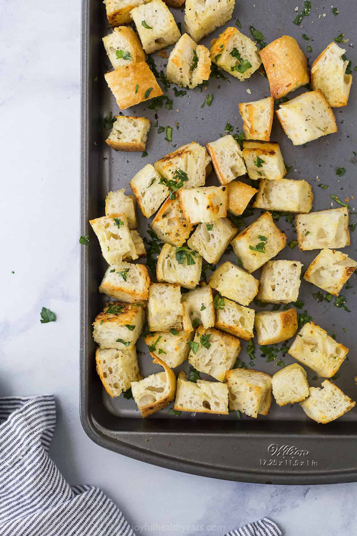 Homemade croutons on a nonstick baking pan with a striped kitchen towel beside it