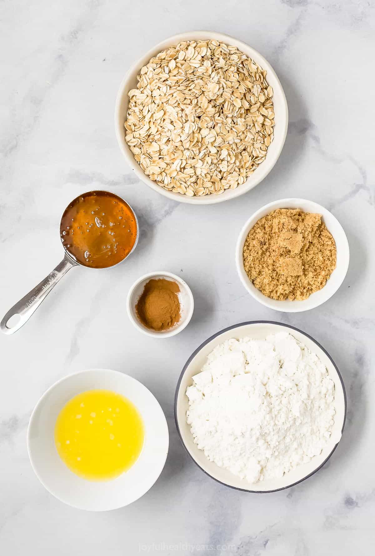 A bowl of rolled oats, a small dish containing ground cinnamon and the rest of the bar ingredients on a marble countertop