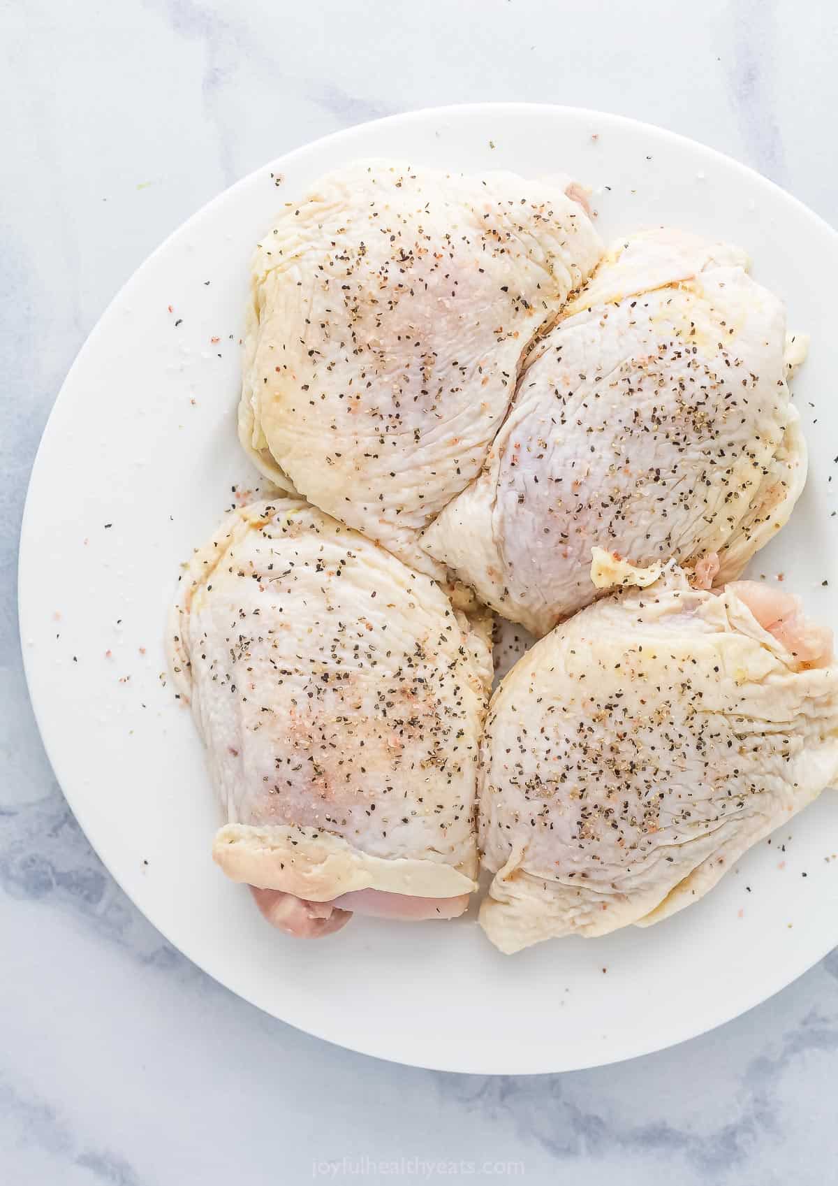 Four seasoned raw chicken thighs on a plate on top of a marble counter