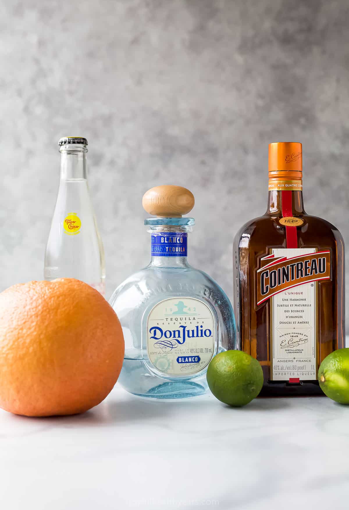 A bottle of cointreau, a bottle of tequila blanco, a bottle of grapefruit sparkling water, a fresh grapefruit and two fresh limes on a marble countertop