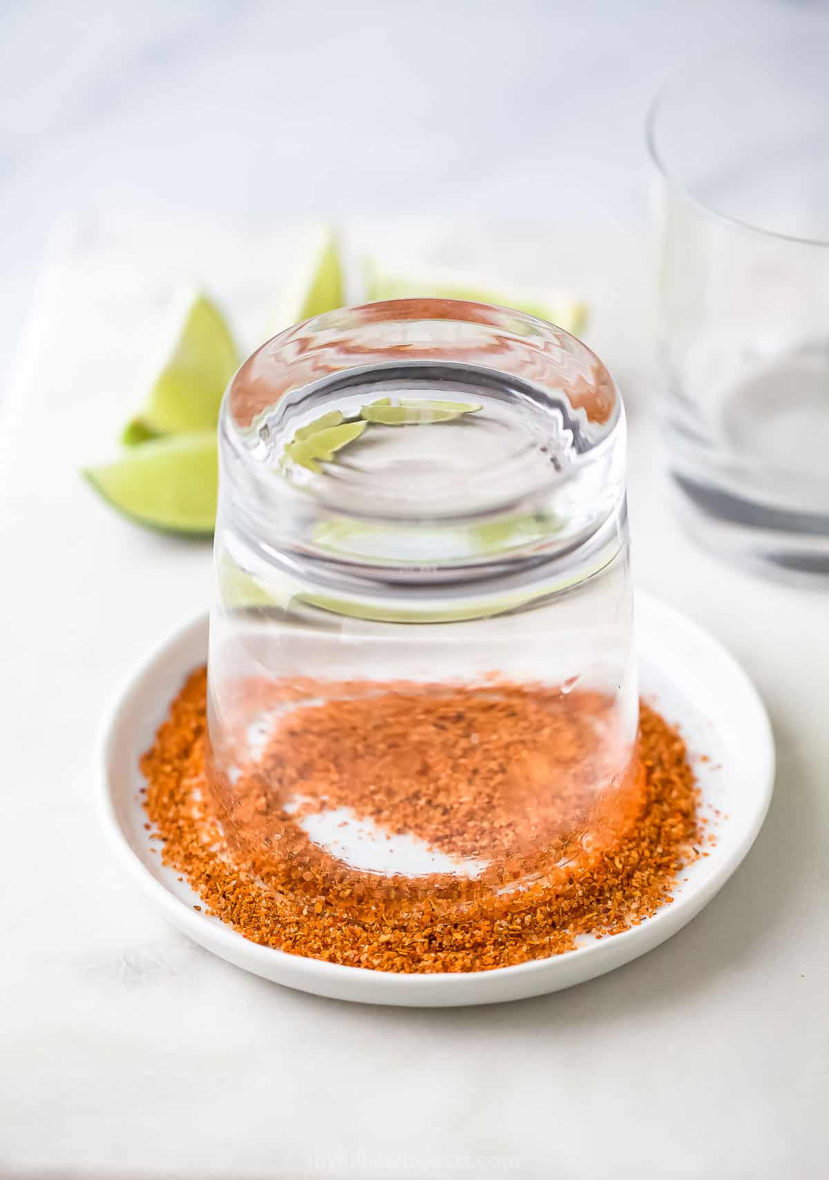 A cocktail glass set rim-down on a plate full of tajín seasoning with fresh lime wedges in the background