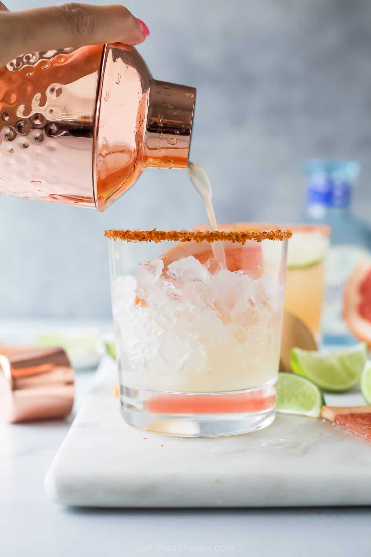 addison: A Paloma cocktail with large clear ice cubes in a tall collins  glass garnished with a grapefruit peel on a white marble counter top