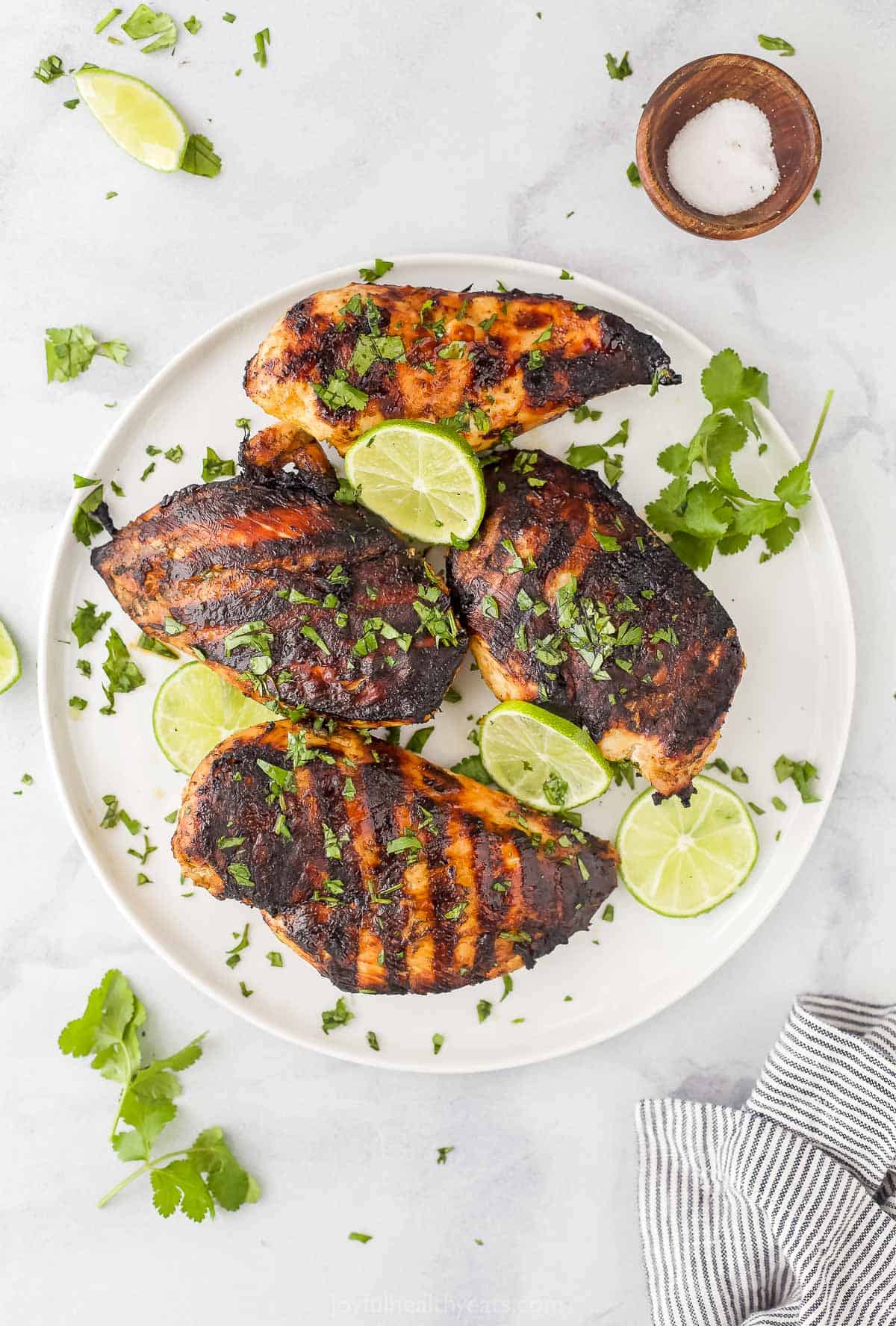 A plate of grilled chicken breasts on a marble counter with a dishtowel and a small dish of salt