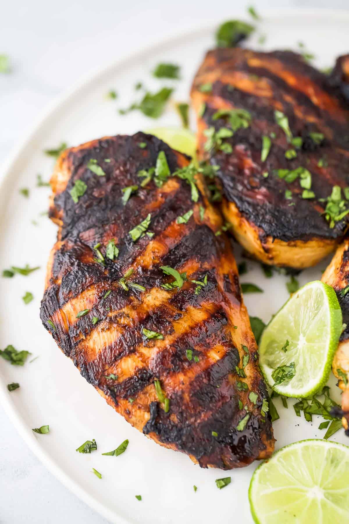 A grilled chicken breast on a plate with lime slices and chopped fresh herbs