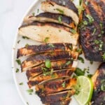 A grilled chicken breast cut into ten slices on a plate with two whole chicken breasts