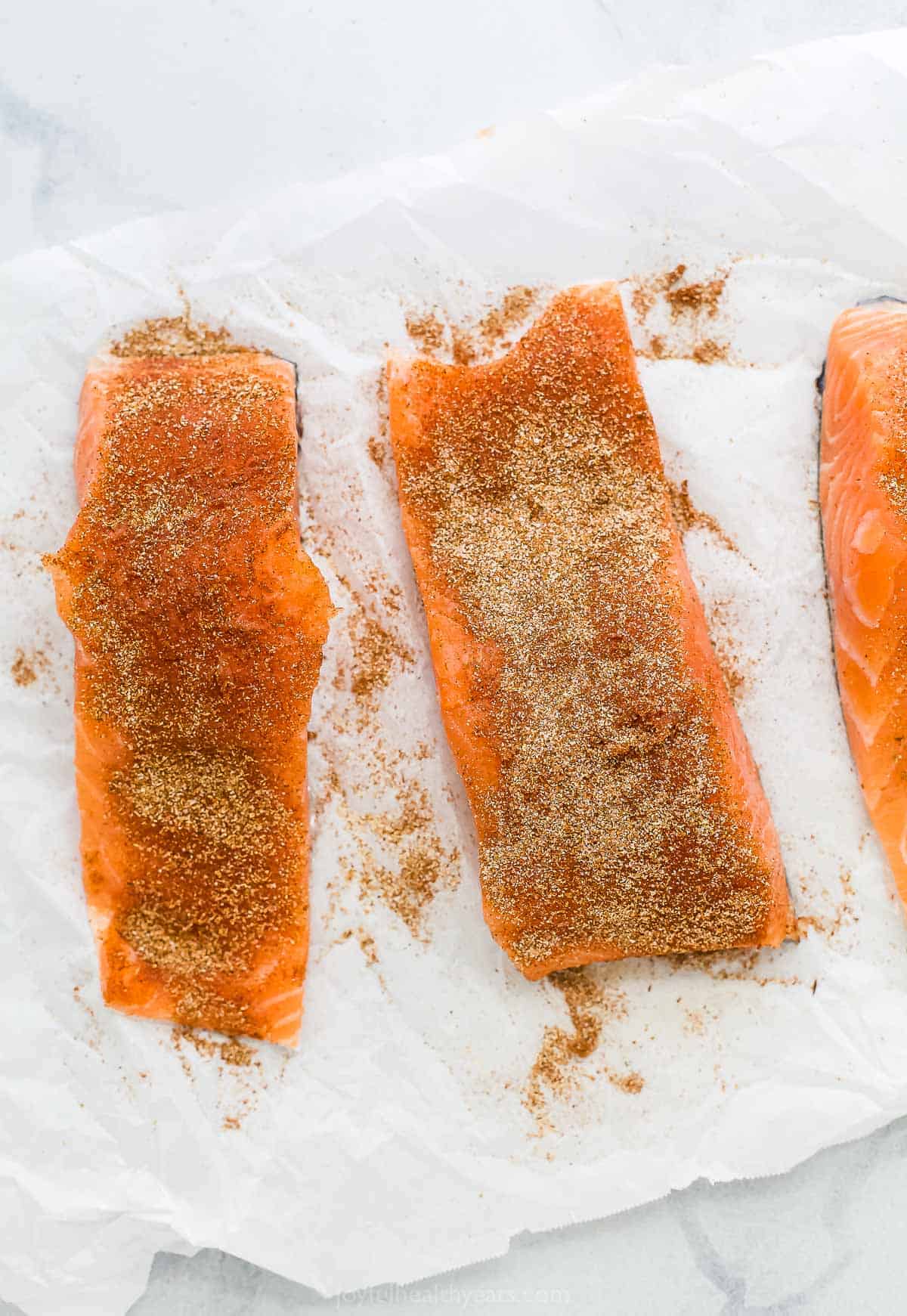 Three seasoned fish fillets lined up on a sheet of parchment paper.