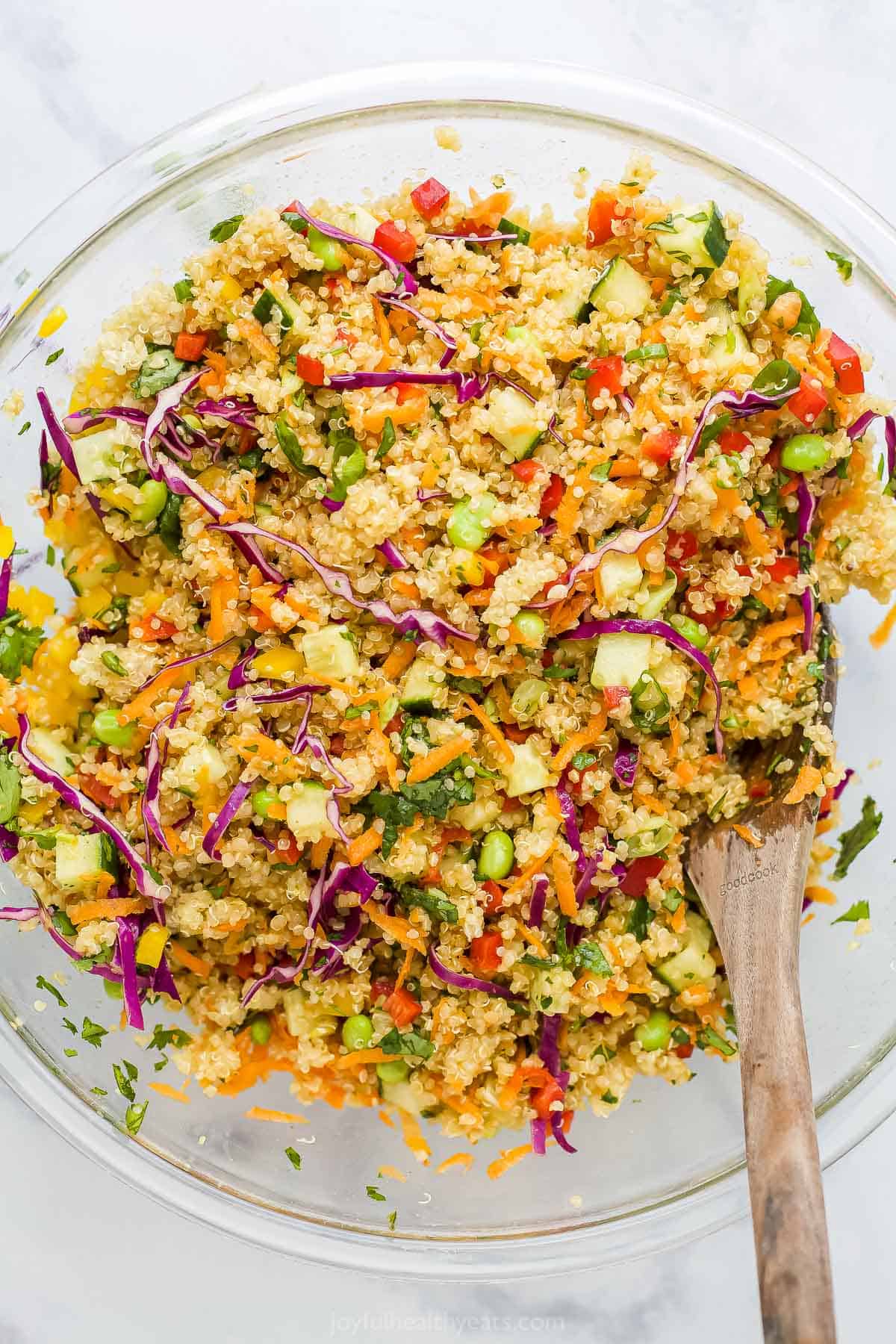 A wooden spoon being used to toss a quinoa salad inside of a large glass mixing bowl