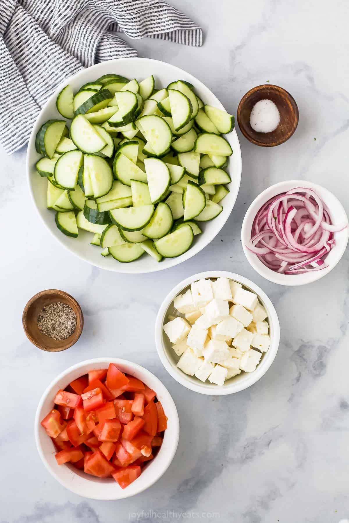 A bowl of chopped tomatoes, a dish full of feta cheese and the rest of the salad ingredients on a kitchen countertop