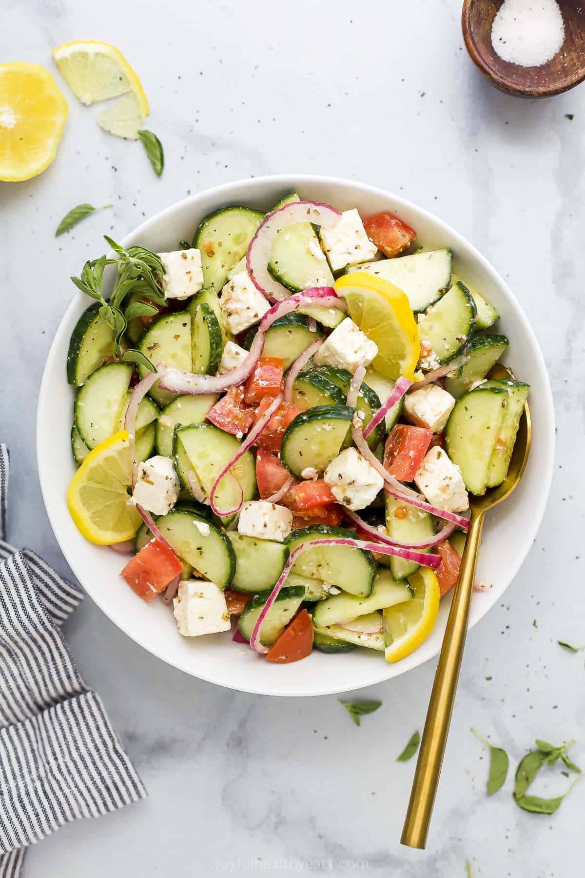 A bowl of cucumber salad on a marble countertop with salt, a fresh lemon and a dishtowel