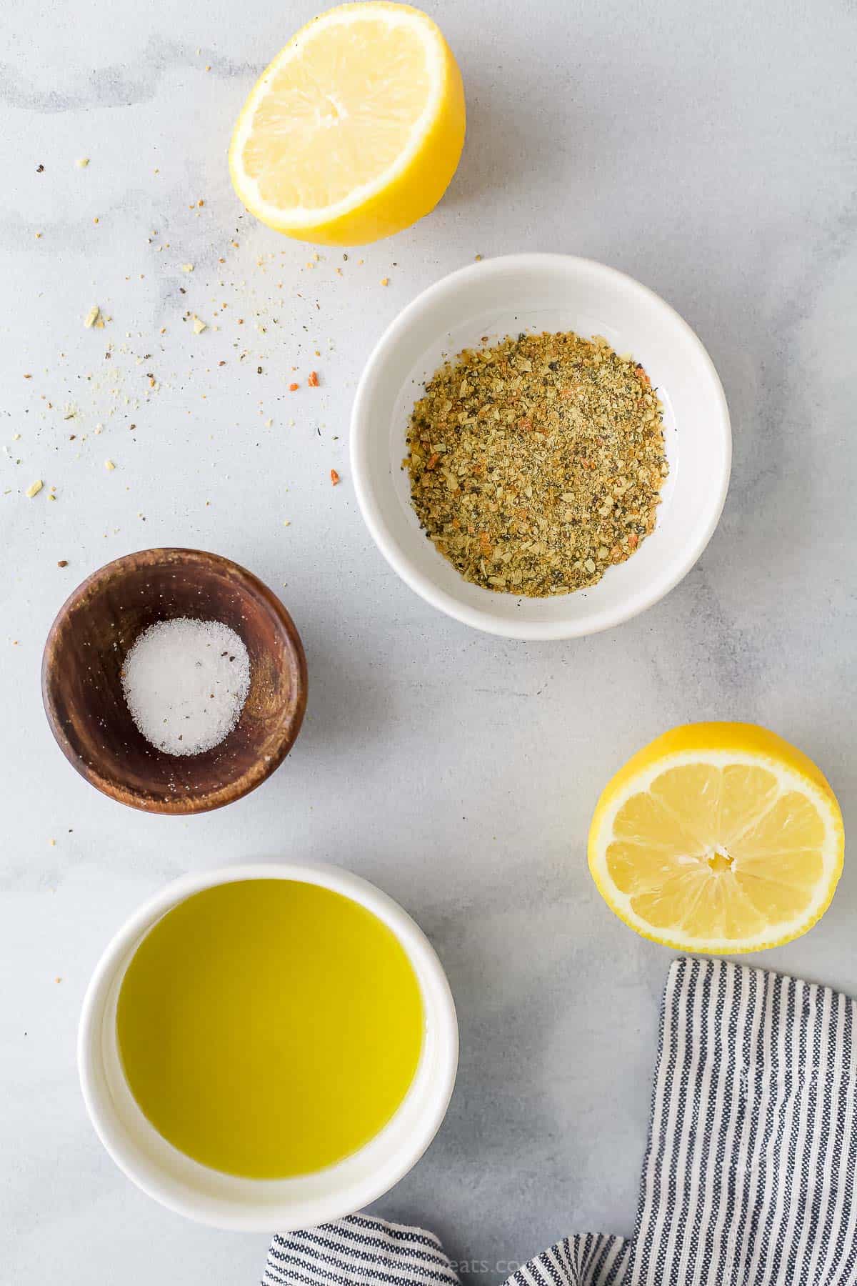 Olive oil, fresh lemon halves and the rest of the ingredients on a kitchen countertop beside a striped dishtowel