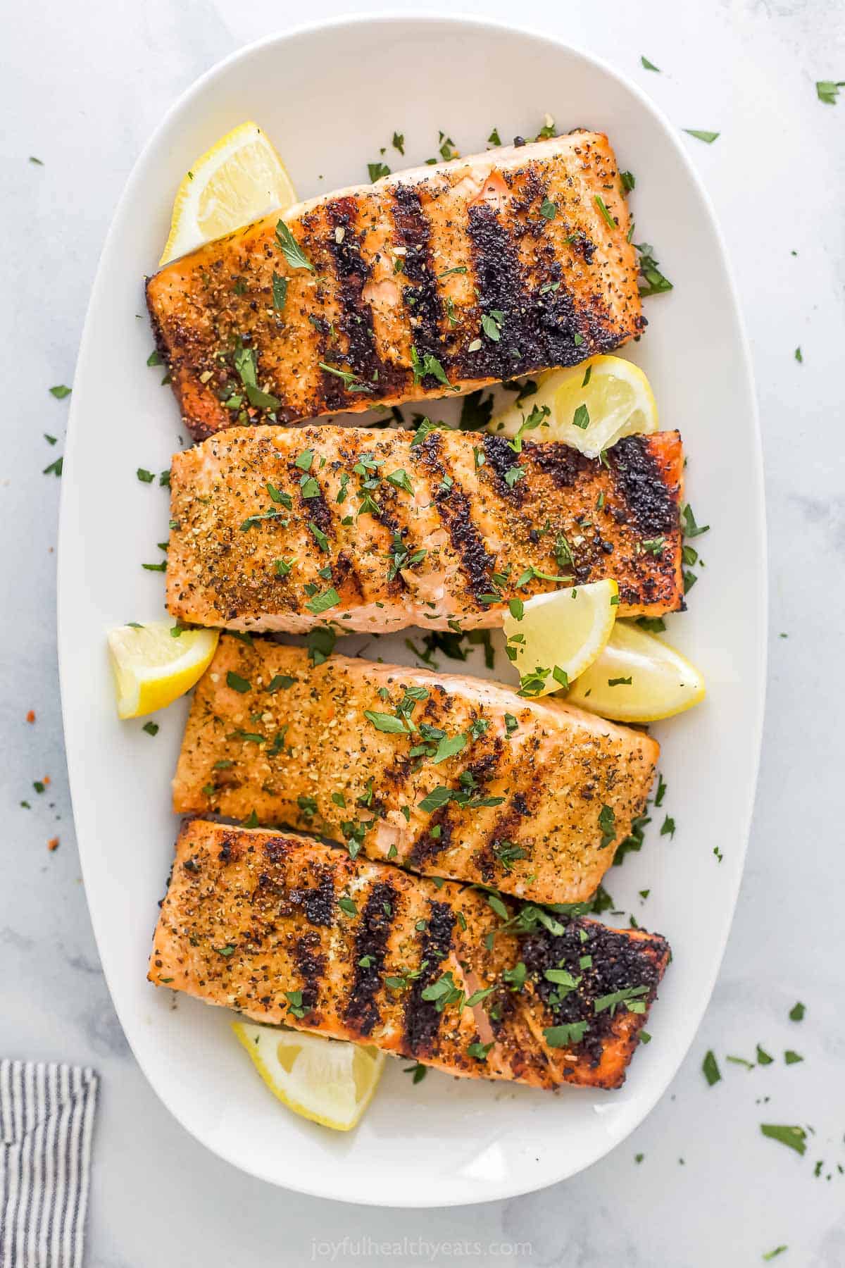 Four lemon pepper grilled salmon fillets lined up on a platter with fresh lemon wedges.