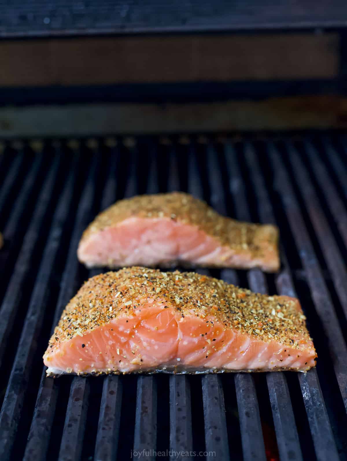 Two raw salmon fillets on a grill with lemon pepper seasoning coating both sides of each fillet.