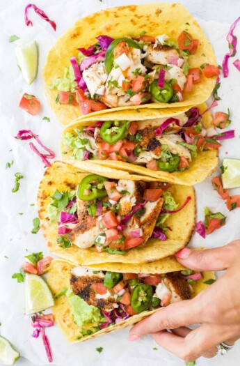 Four mahi mahi tacos lined up on a kitchen countertop with a hand grabbing one from the end of the line