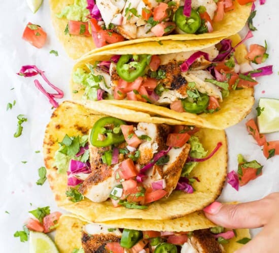 Four mahi mahi tacos lined up on a kitchen countertop with a hand grabbing one from the end of the line