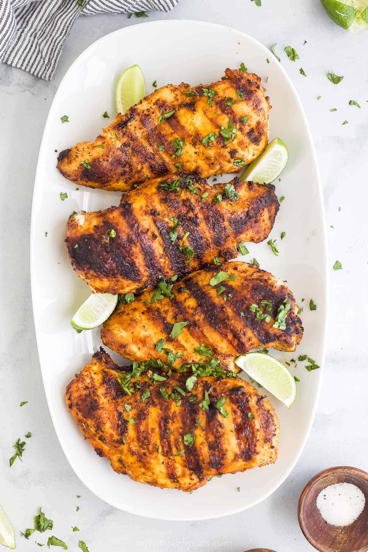 Four grilled chicken breasts on a serving platter with a kitchen towel and a dish of salt beside it