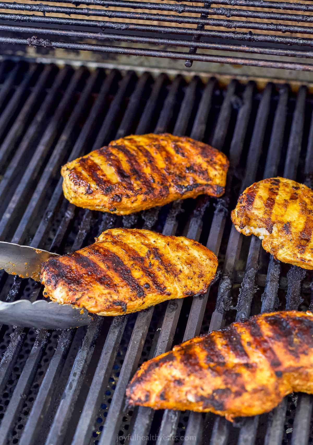 Four chicken breasts searing on a grill with a pair of tongs grasping one of them