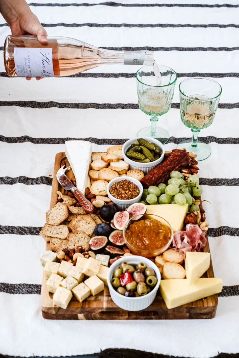 Wine is poured into glasses next to a charcuterie board filled with food items.