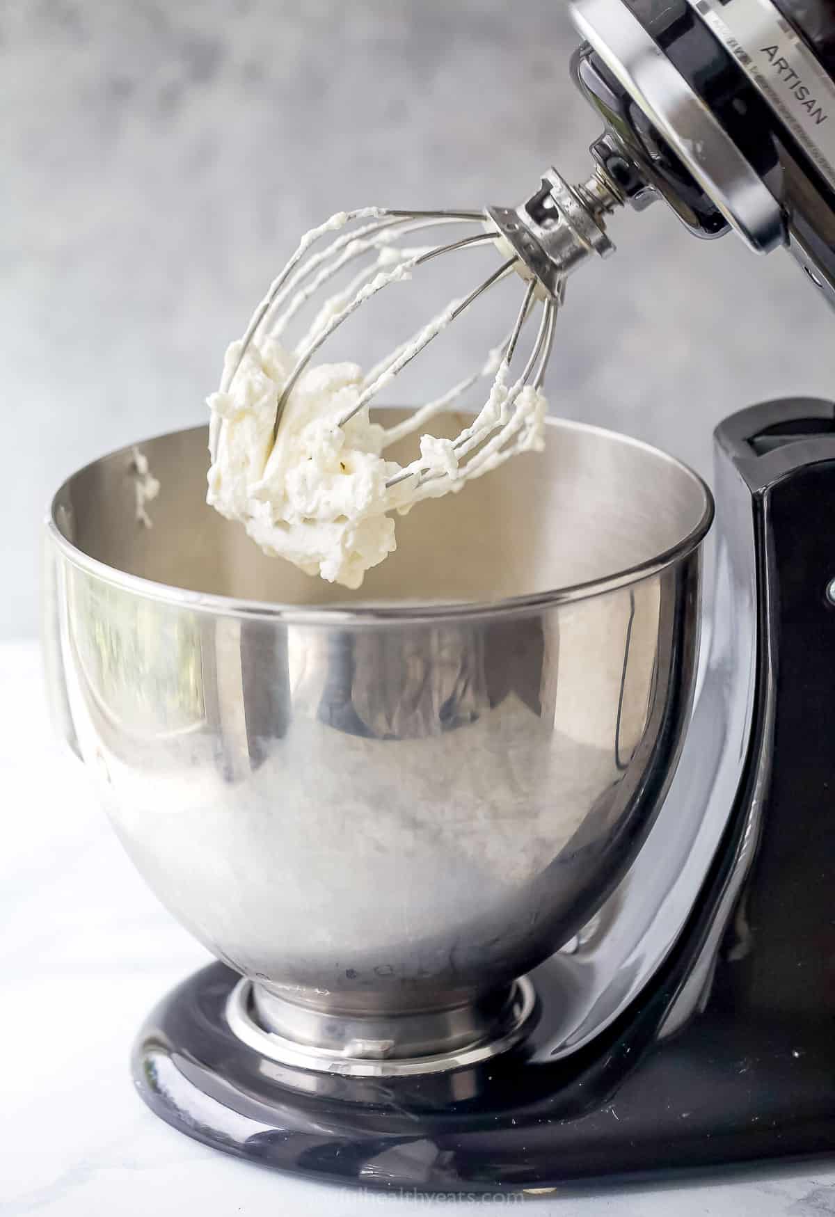 An electric mixer fitted with a large metal bowl and a whisk attachment with whipped cream being beaten