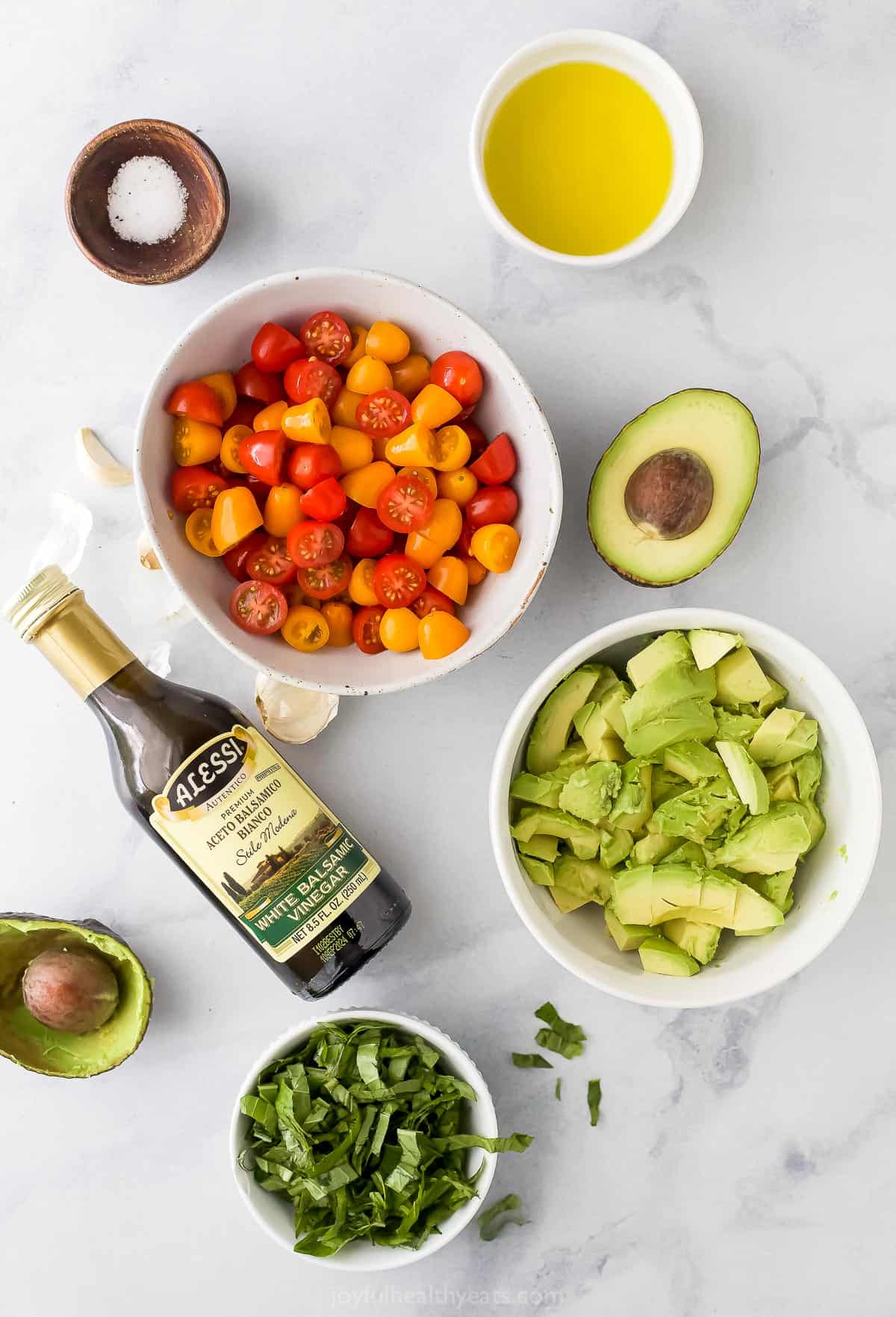 A bottle of olive oil, a dish of white balsamic vinegar, a bowl of fresh basil and the rest of the topping ingredients on a marble countertop