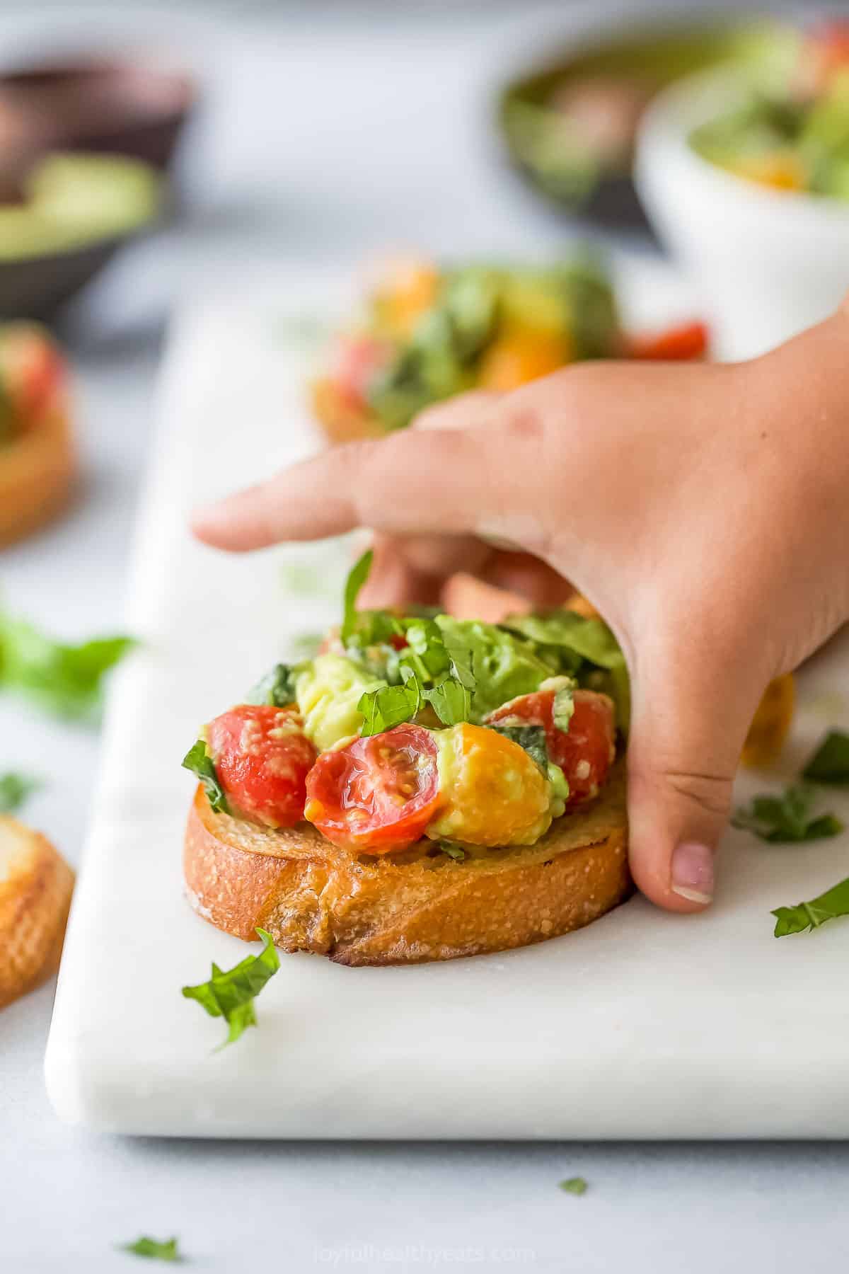 A slice of avocado bruschetta on a cutting board with a hand in the process of grabbing it
