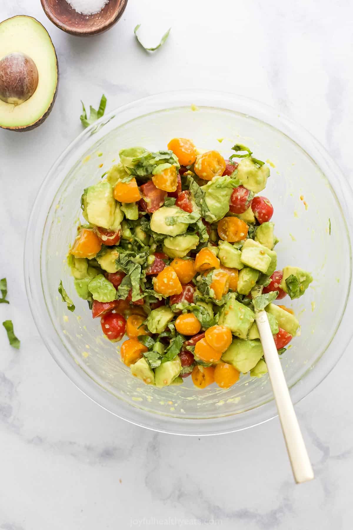 A bowl full of bruschetta topping with a halved avocado and a dish of salt beside it