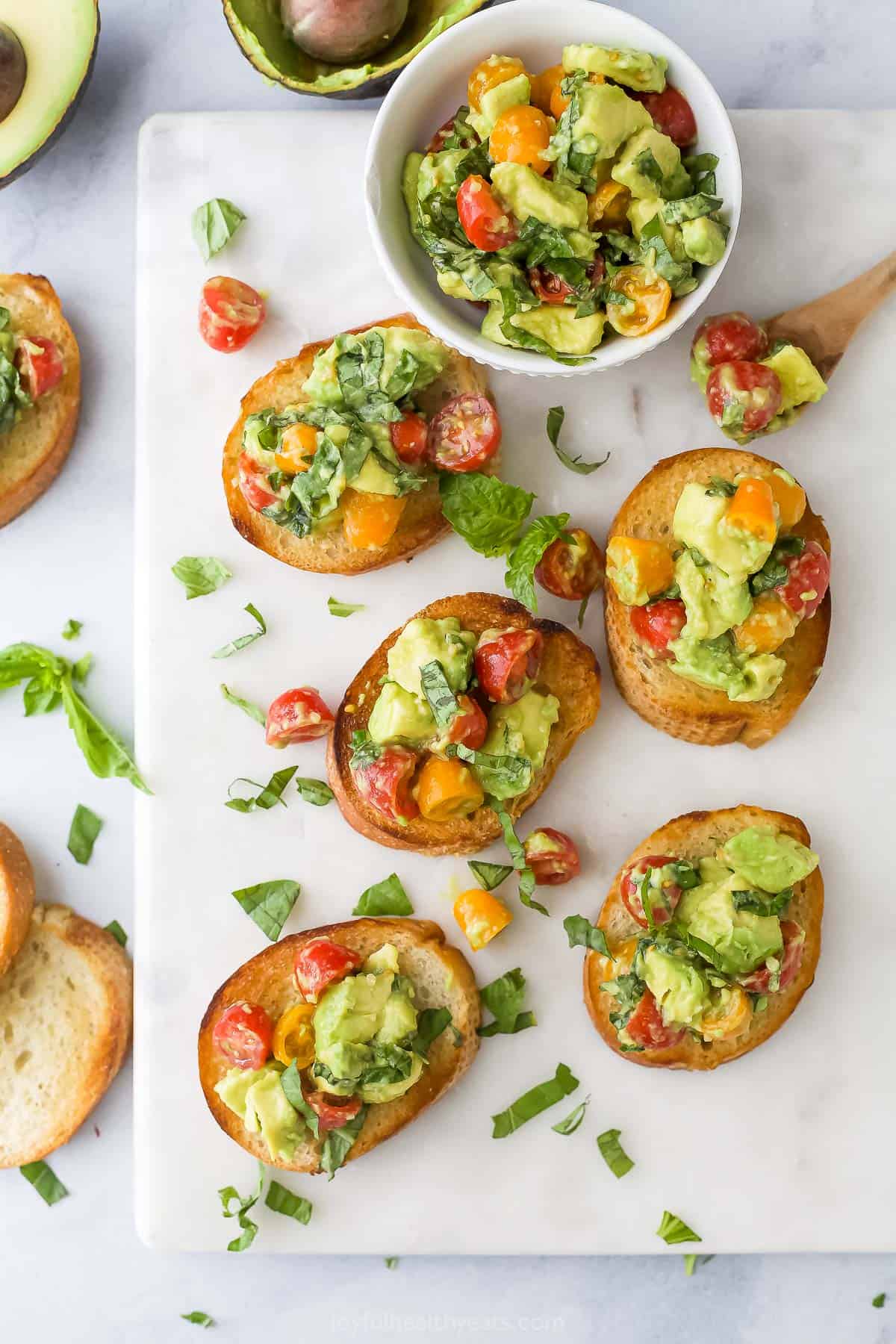 Five pieces of toast topped with avocado, tomatoes, basil, olive oil and white balsamic vinegar