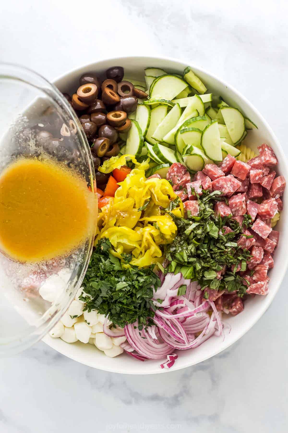 Italian vinaigrette dressing being poured over a bowl of veggies, herbs, salami, cheese and pasta