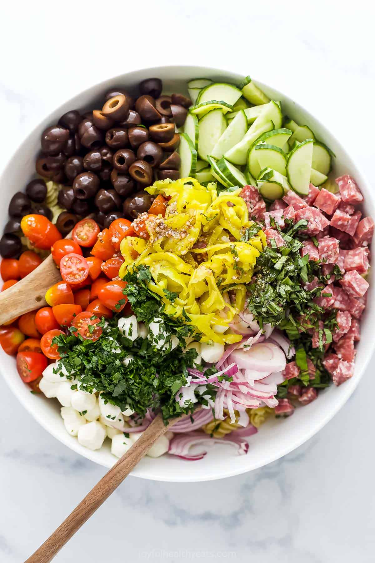 Vegetables, pasta, herbs, salami, mozzarella and vinaigrette being mixed together in a big bowl