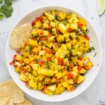 A bowl of mango salsa on a marble countertop with a few tortilla chips beside it