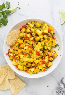 A bowl of mango salsa on a marble countertop with a few tortilla chips beside it