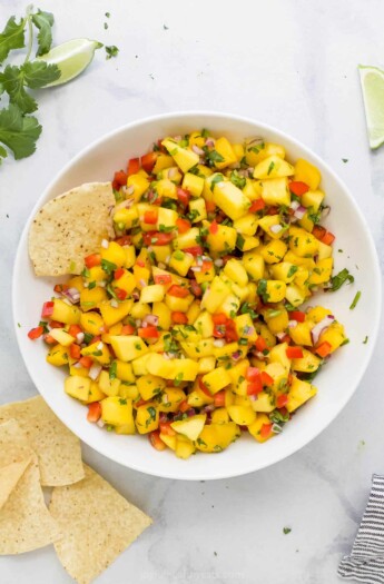 A bowl of mango salsa on a marble countertop with a few tortilla chips beside it