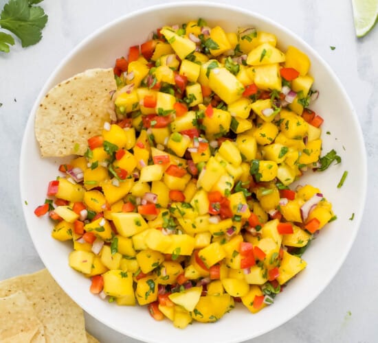 A bowl of mango salsa on a marble countertop with a few tortilla chips beside it