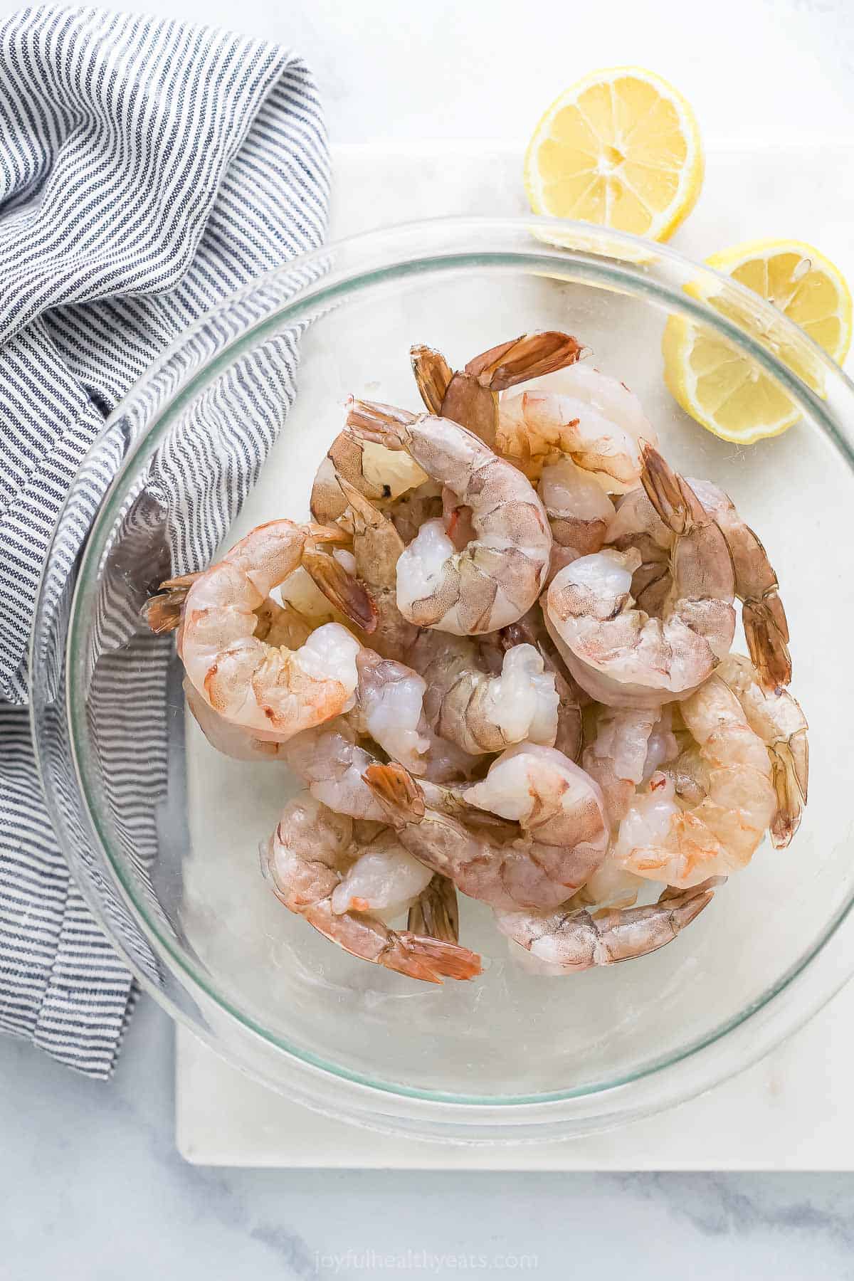 Peeled and deveined shrimp inside of a glass bowl next to a halved lemon and a cloth napkin